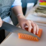 Close-up of a chef's hands slicing fresh salmon for sushi on a cutting board indoors.